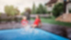 This image shows two young children sitting on the edge of a pool with their feet in the water, laughing and splashing